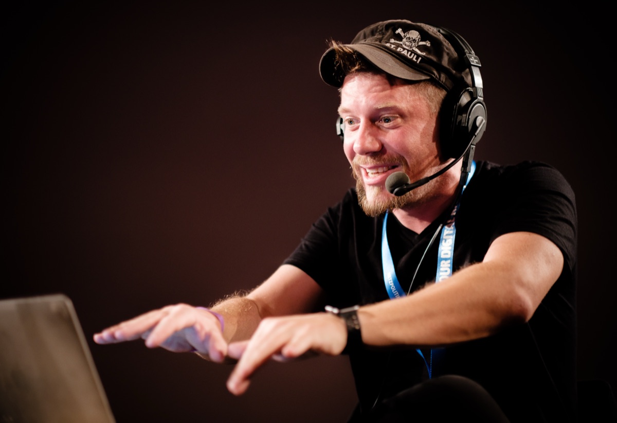 Linus Neumann mit St.-Pauli-Cap und Headset gestikuliert lachend vor einem Laptop auf einer Bühne