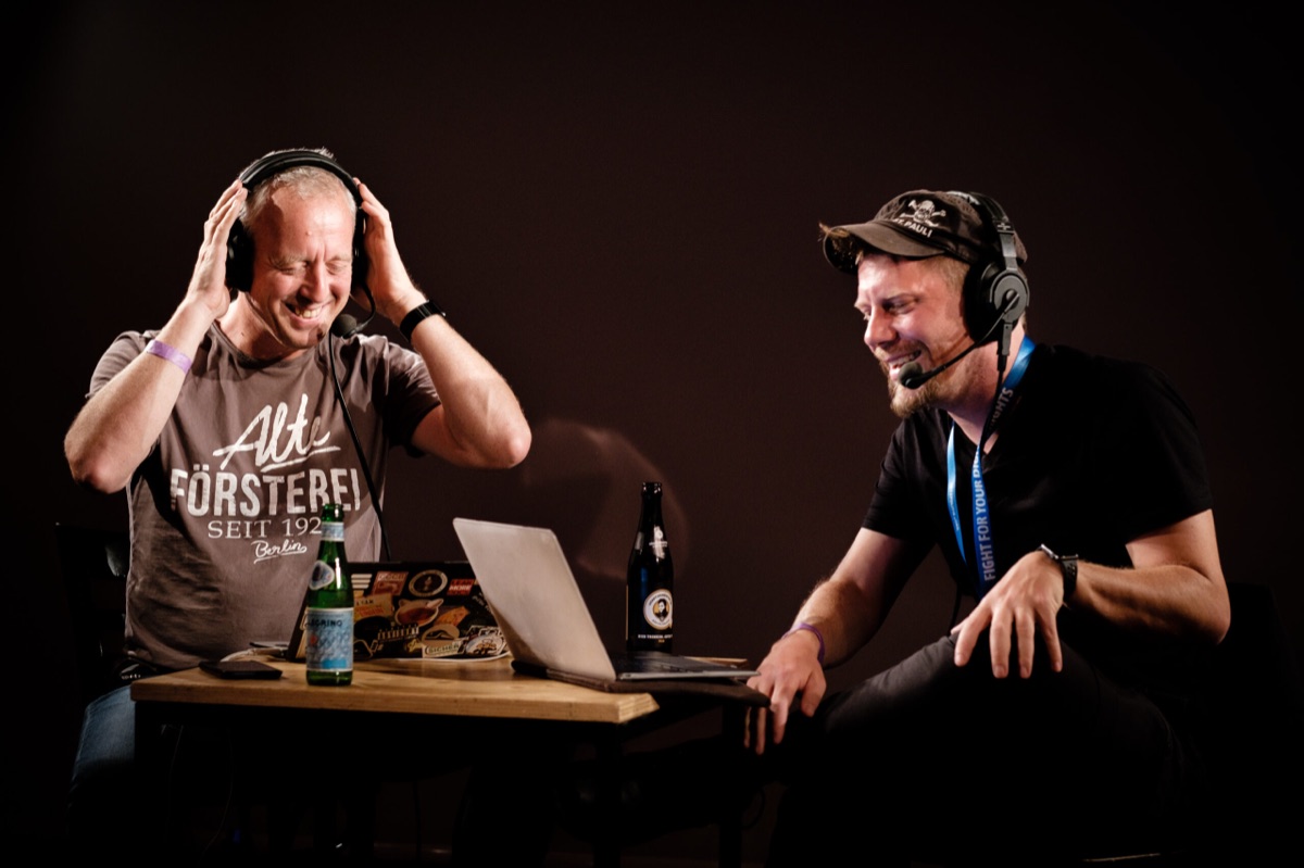 Tim Pritlove und Linus Neumann mit Headsets lachen bei einer Live-Aufnahme des Podcasts an einem Tisch mit Laptop und Getränken